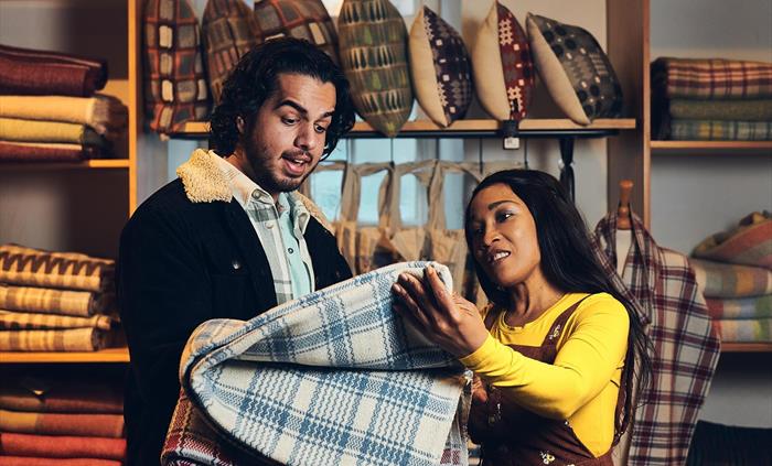A couple stand in the museum shop. They are holding up a blue and white check blanket, to buy and take home. The blanket was made on the looms at the